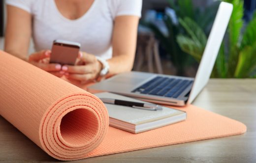 agile BGF » betriebliche Gesundheitsförderung » arbeitende Person mit Laptop, Handy, Block und Stift sowie Fitnessmatte auf dem Tisch